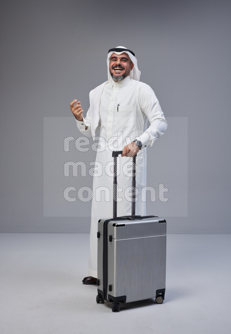 Saudi man wearing Thob and white Shomag standing holding Travel bag on Gray background