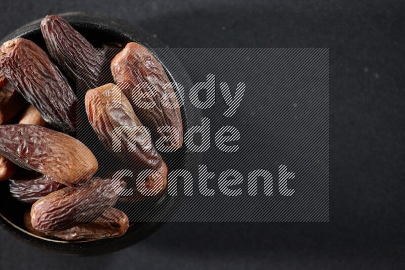 A black pottery bowl full of dried dates on a black background in different angles