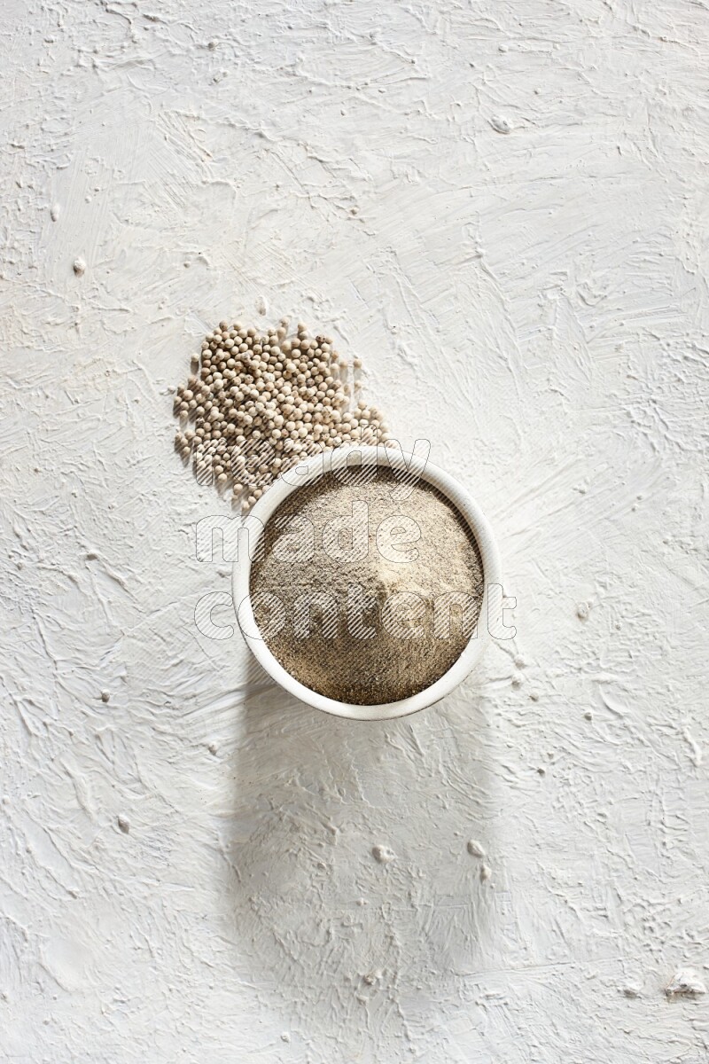 وعاء فخاري أبيض ممتلئ ببودرة الفلفل الأبيض مع حبوب الفلفل على أرضية بيضاء