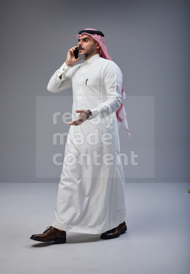 Saudi man Wearing Thob and red Shomag standing talking on phone on Gray background