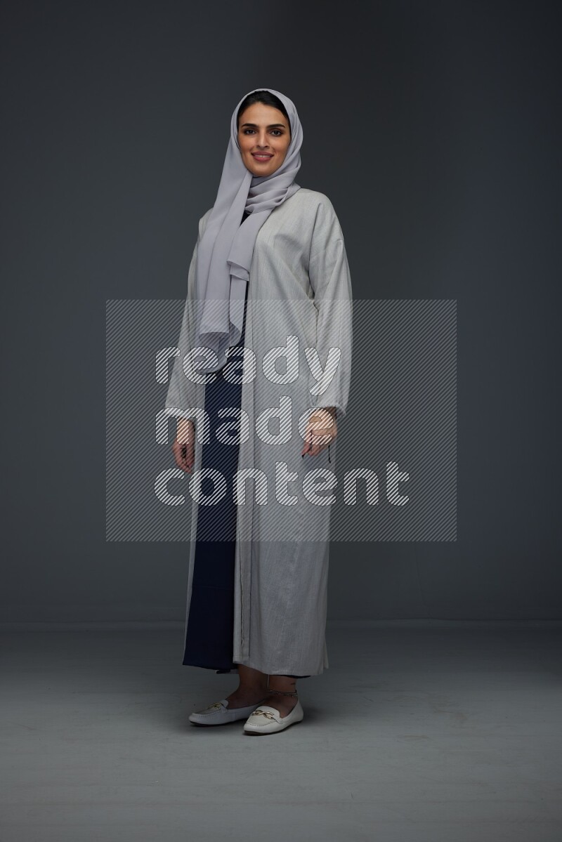 A Saudi woman wearing a light gray Abaya and head scarf standing and making multi poses eye level on a grey background