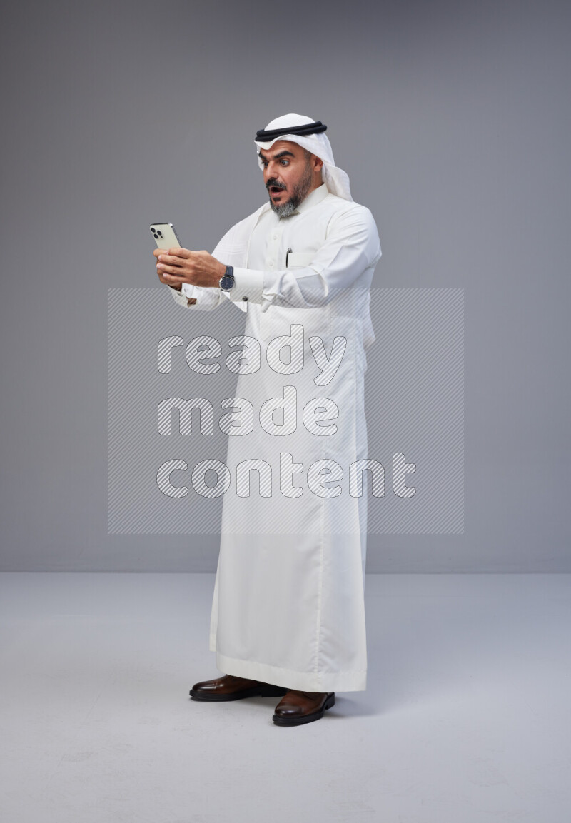 Saudi man Wearing Thob and white Shomag standing texting on phone on Gray background