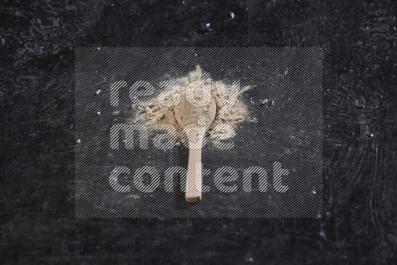 A wooden spoon full of garlic powder with scattered powder around it on a textured black flooring