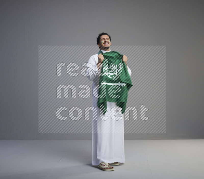 A Saudi man standing wearing thob with face painting wrapping Saudi flag scarf and holding big Saudi flag on gray background