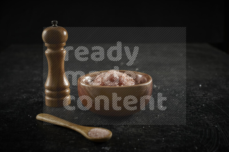 A wooden bowl and spoon filled with coarse pink himalayan salt and a wooden grinder beside them on black background