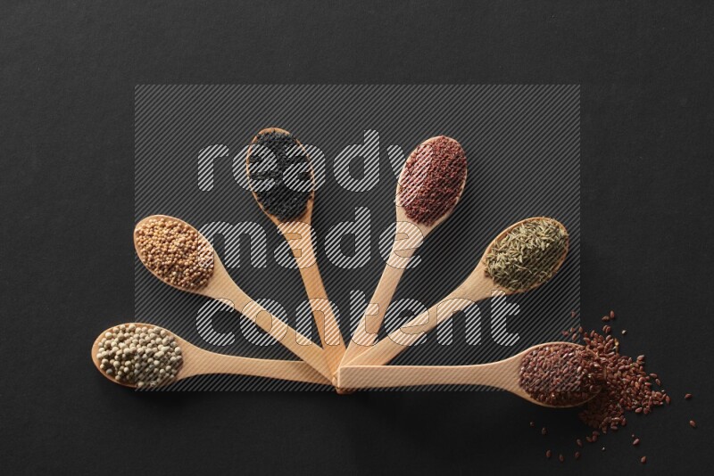 wooden spoons filled with white peppers, mustard seeds, black seeds, garden cress, cumin and flax on black flooring