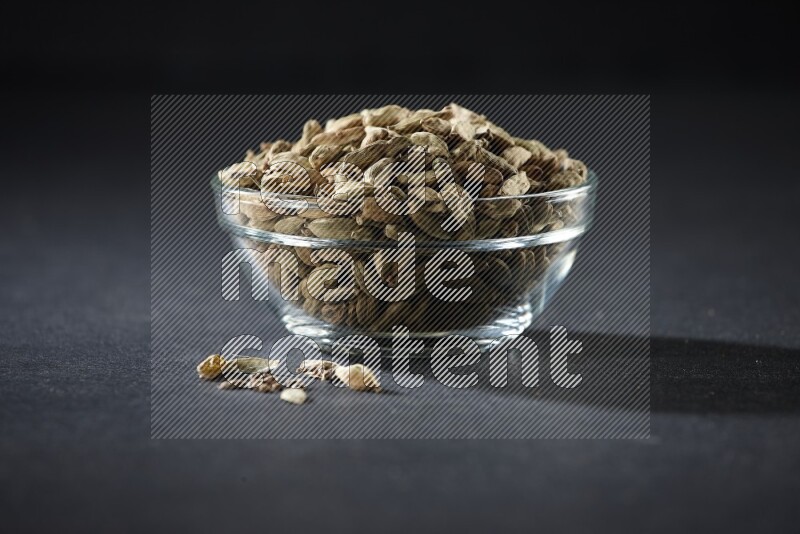 A glass bowl full of cardamom and more seeds spreaded beneath the bowl on black flooring