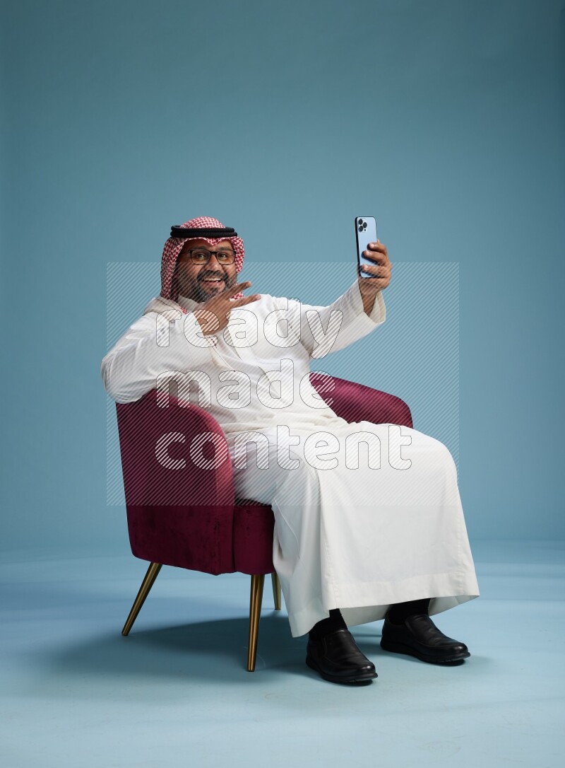 Saudi Man with shimag sitting on chair taking selfie on blue background