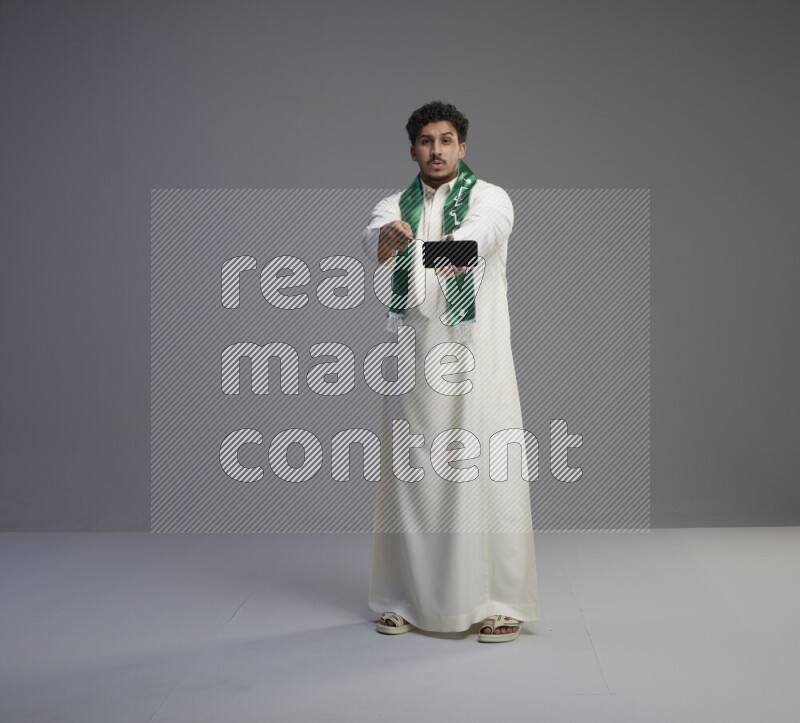 A Saudi man standing wearing thob and Saudi flag scarf showing phone to camera on gray background