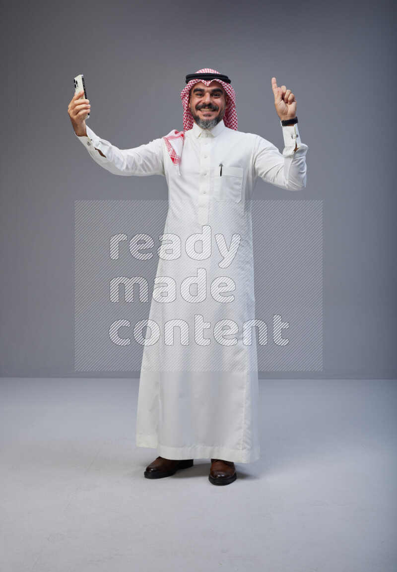 Saudi man Wearing Thob and red Shomag standing taking selfie on Gray background