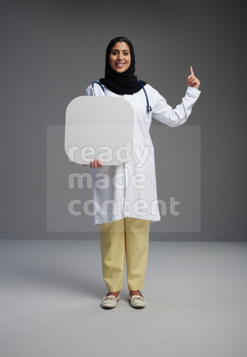 Saudi woman wearing lab coat with stethoscope standing holding social media sign on Gray background