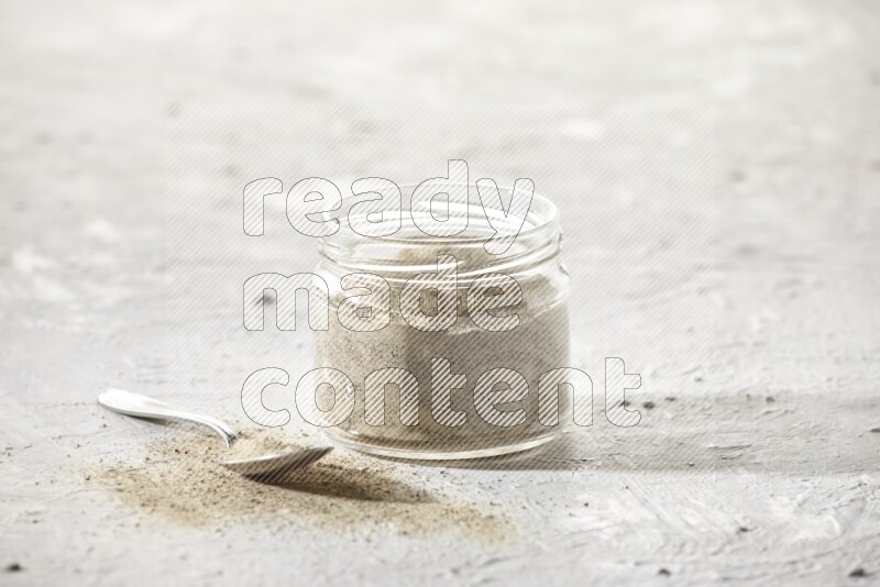 A glass jar and a metal spoon full of white pepper powder on textured white flooring