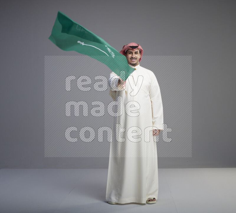 A Saudi man standing wearing thob and red shomag raising big Saudi flag on gray background