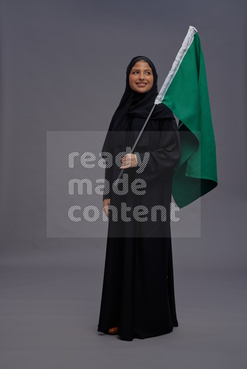 Saudi woman wearing Abaya standing holding Saudi flag on gray background