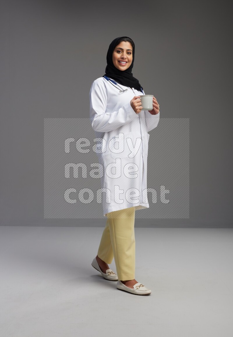 Saudi woman wearing lab coat with stethoscope standing holding mug on Gray background