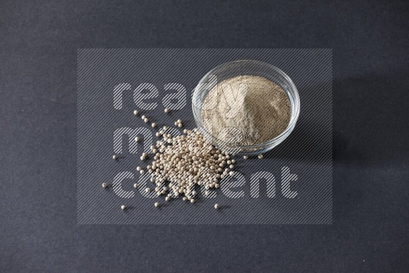 A glass bowl full of white pepper powder with white pepper beads on black flooring