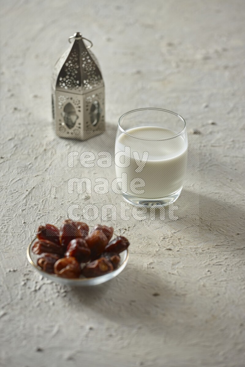 A silver lantern with different drinks, dates, nuts, prayer beads and quran on textured white background