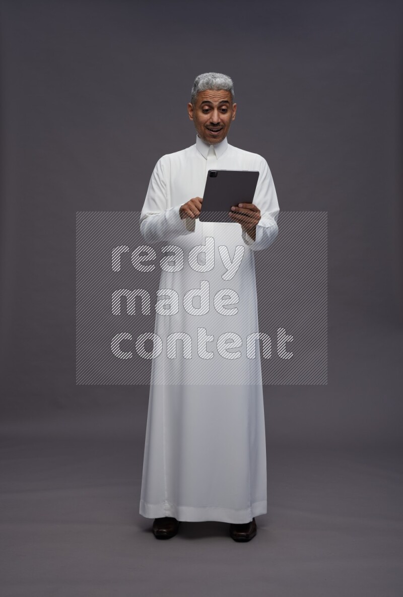 Saudi man wearing thob standing working on tablet on gray background