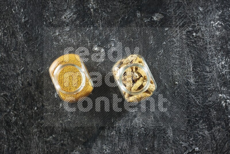 وعاءان زجاجيان للتوابل ممتلئان ببودرة الكركم وأصابع الكركم المجففة على خلفية سوداء