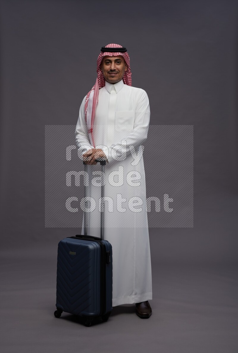 Saudi man wearing thob and shomag standing holding bag on gray background