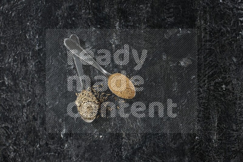 2 metal spoons full of cumin seeds and cumin powder on a textured black flooring