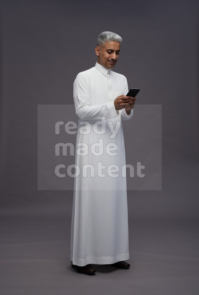 Saudi man wearing thob standing texting on phone on gray background
