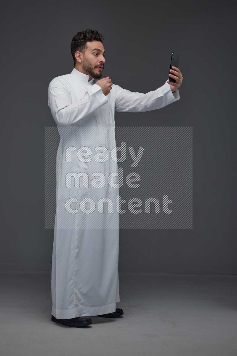 A Saudi man wearing Thobe and taking selfie with his phone making different poses eye level on a gray background