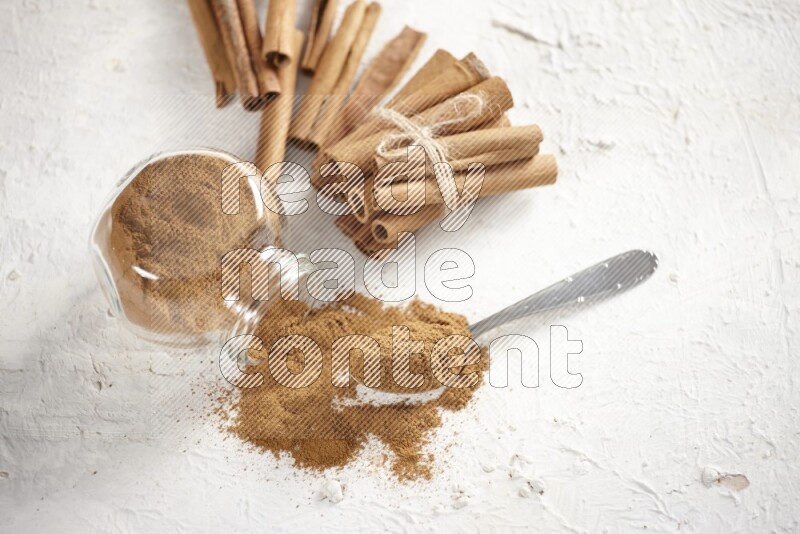 Flipped herbs glass jar full of cinnamon powder with a metal spoon full of powder and cinnamon sticks on a textured white background