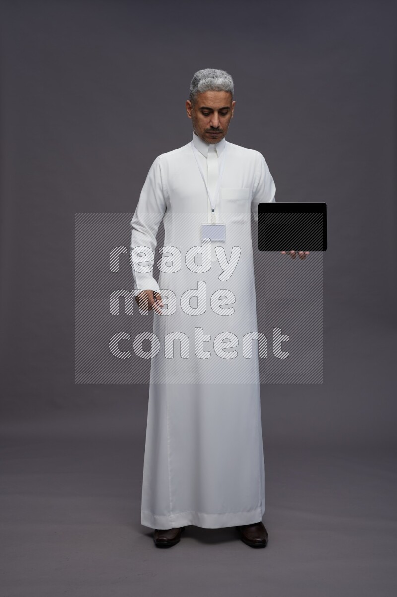 Saudi man wearing thob with neck strap employee badge standing showing tablet to camera on gray background