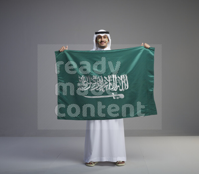 A Saudi man standing wearing thob and white shomag with face painting holding big Saudi flag on gray background