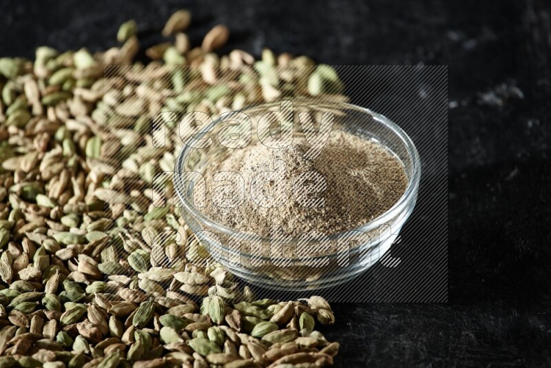 A glass bowl full of cardamom powder and cardamom seeds spreaded beneath the bowl on textured black flooring