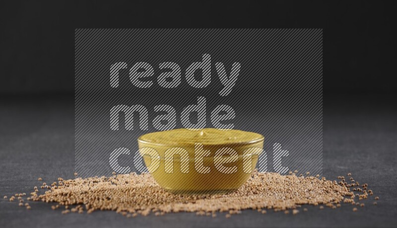 A glass bowl full of mustard paste with mustard seeds underneath on black flooring