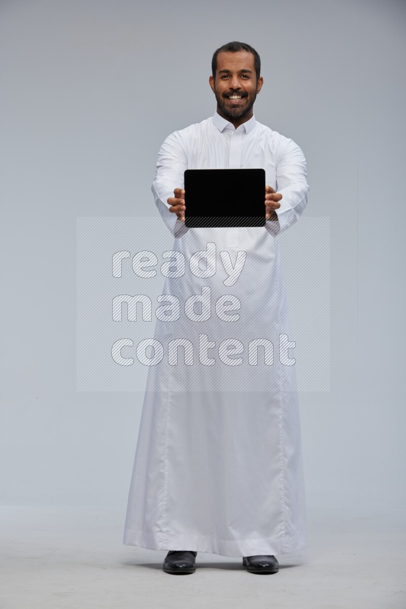 Saudi man wearing Thob standing showing tablet to camera on Gray background