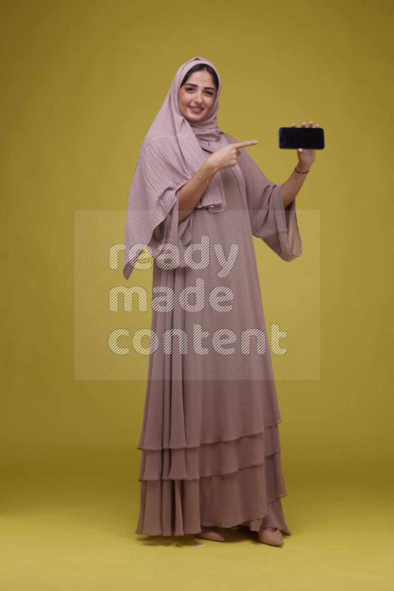 A woman Showing her Phone screen on a Yellow Background wearing Brown Abaya with Hijab