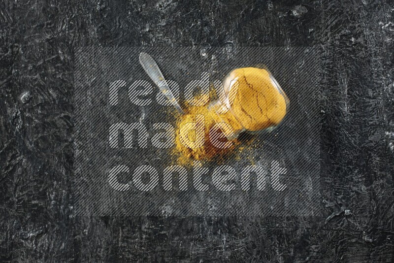 A flipped glass spice jar and a metal spoon full of turmeric powder and powder spilled out of it on textured black flooring
