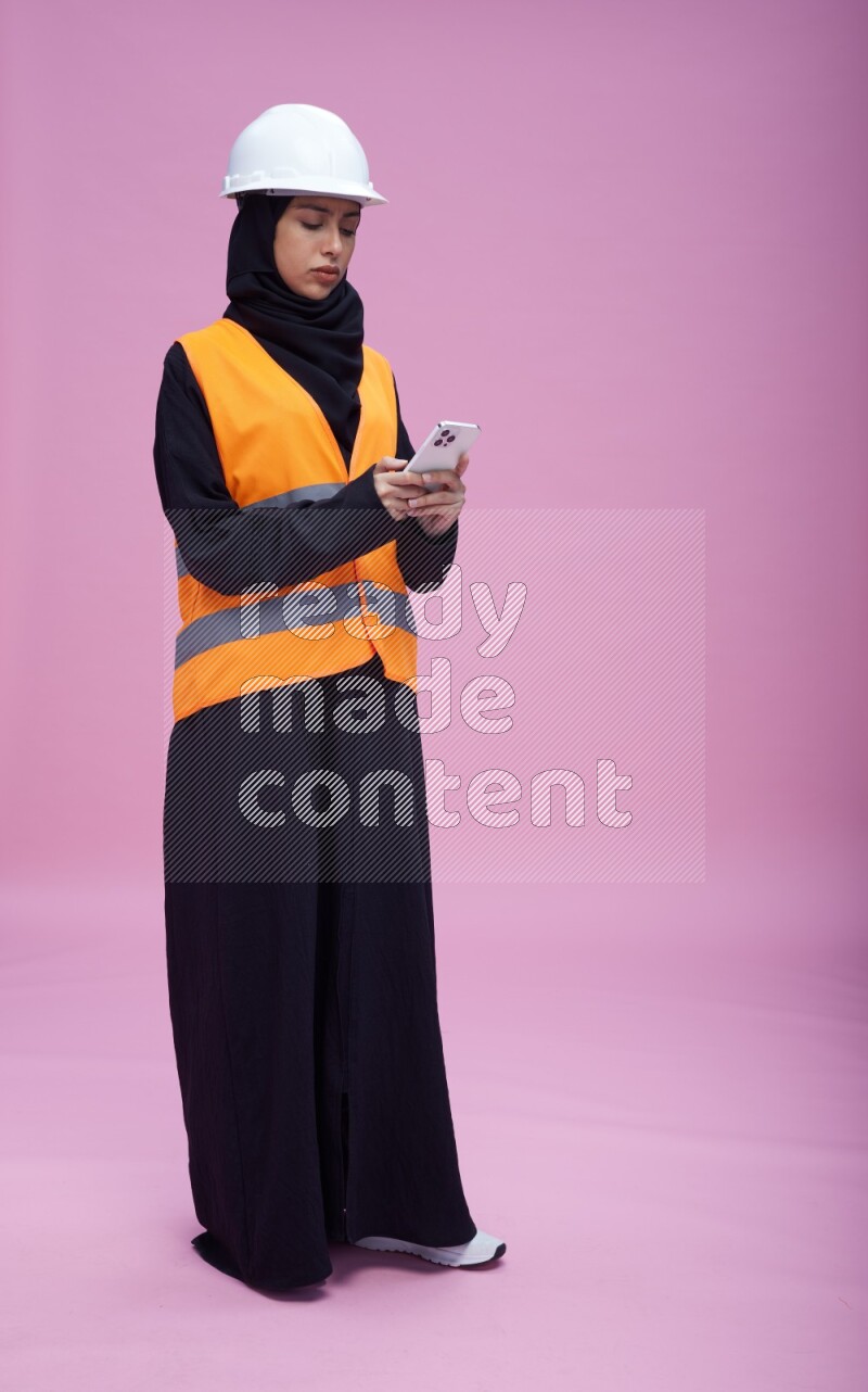 Saudi woman wearing Abaya with engineer vest and helmet standing texting on phone on pink background
