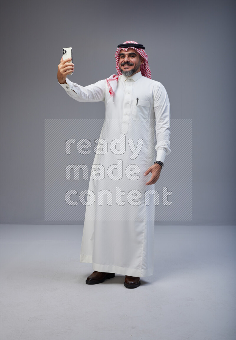 Saudi man Wearing Thob and red Shomag standing taking selfie on Gray background