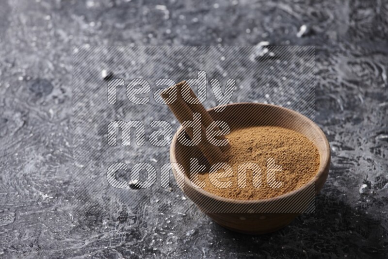 Wooden bowl full of cinnamon powder and a cinnamon stick on a textured black background