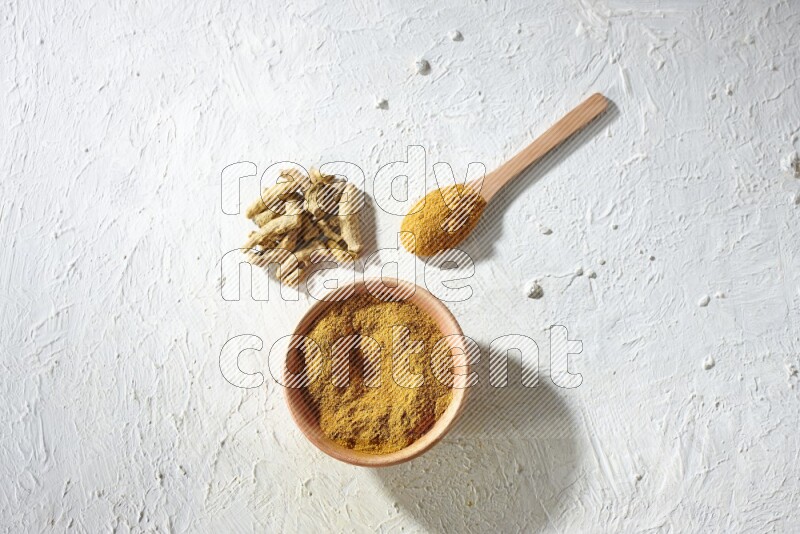 A wooden bowl and wooden spoon full of turmeric powder with dried turmeric fingers on textured white flooring