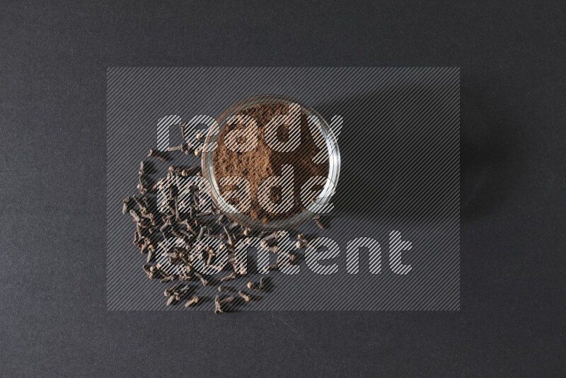 A glass bowl full of cloves powder with cloves grains spread on black flooring