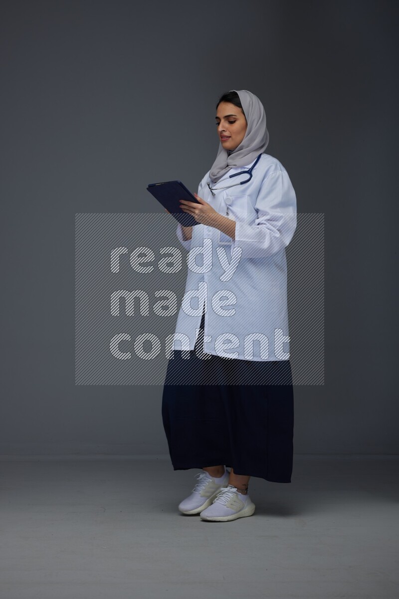 A Saudi doctor wearing a light gray head scarf standing and crossing her hands eye level on a grey background