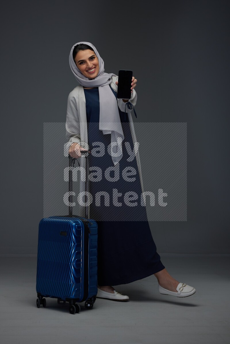 A Saudi woman wearing a light gray Abaya and head scarf standing and showing the phone's screen while wearing headphones eye level on a grey background