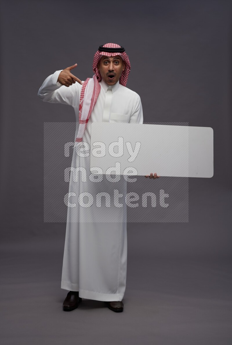 Saudi man wearing thob and shomag standing holding board on gray background