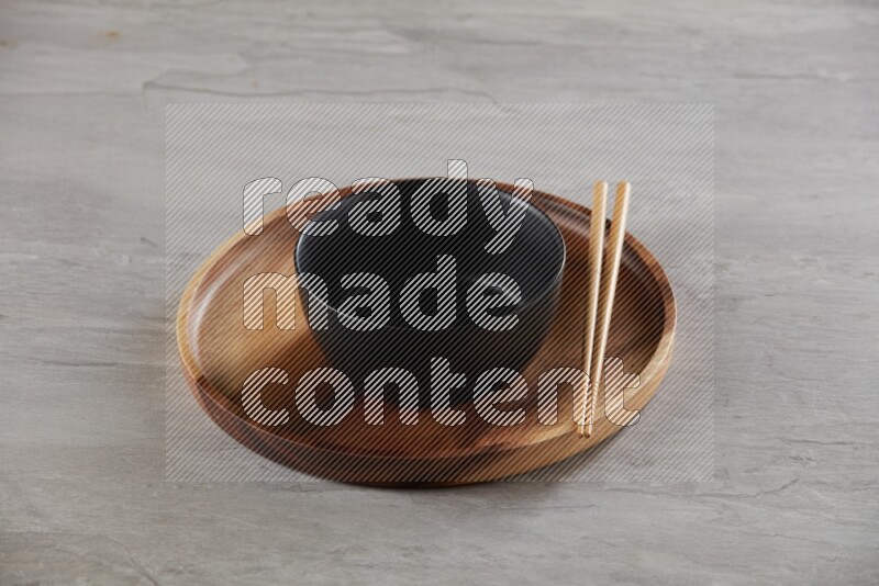 black ceramic round bowl on top of brown wood round plate and wood chopsticks, on grey textured countertop