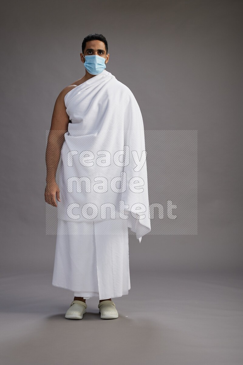 A man wearing Ehram with face mask Standing Interacting with the camera on gray background