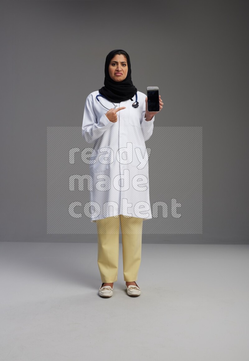 Saudi woman wearing lab coat with stethoscope standing showing phone to camera with sign in the back on Gray background
