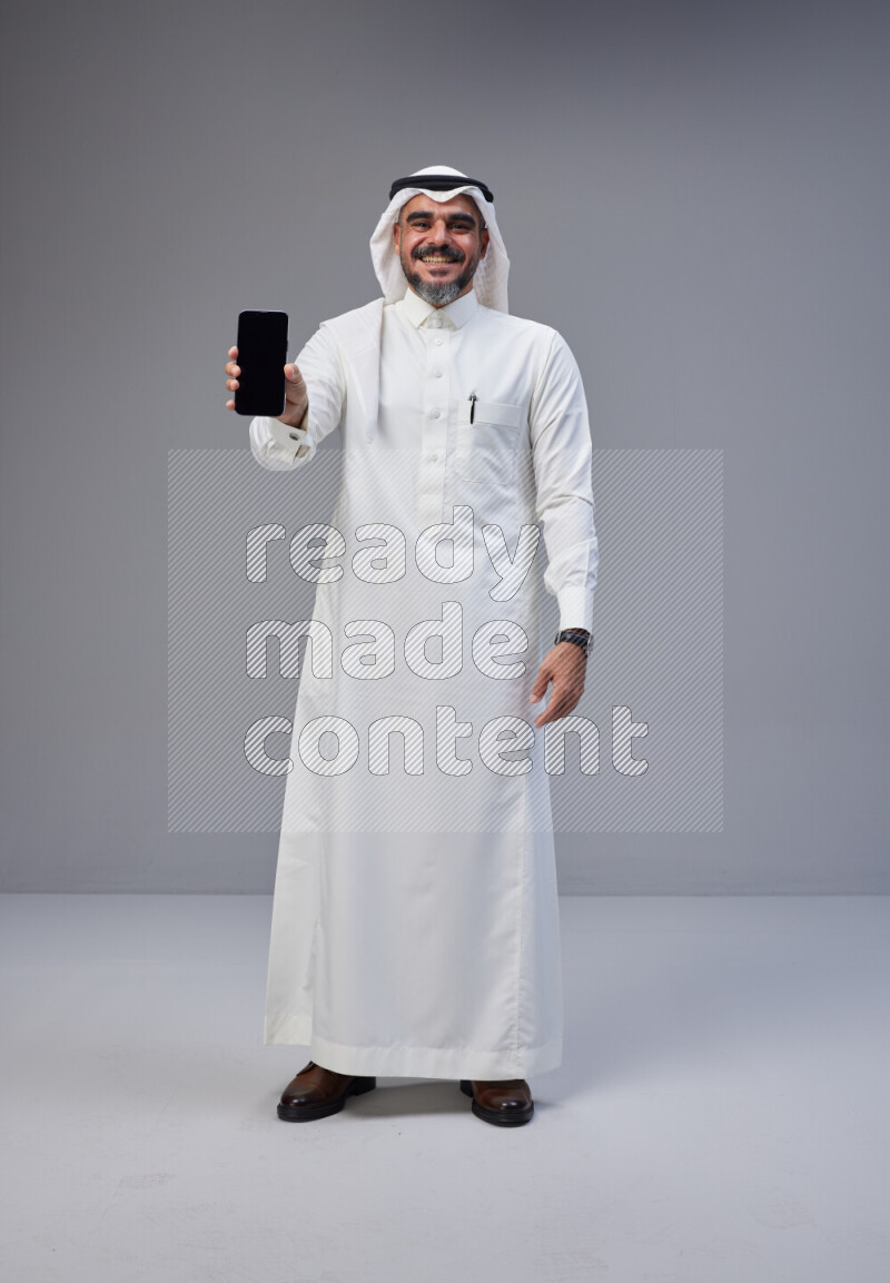 Saudi man Wearing Thob and white Shomag standing showing phone to camera on Gray background