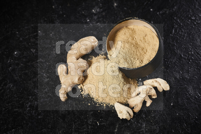 A black pottery bowl full of ground ginger powder with fallen powder from it on black background