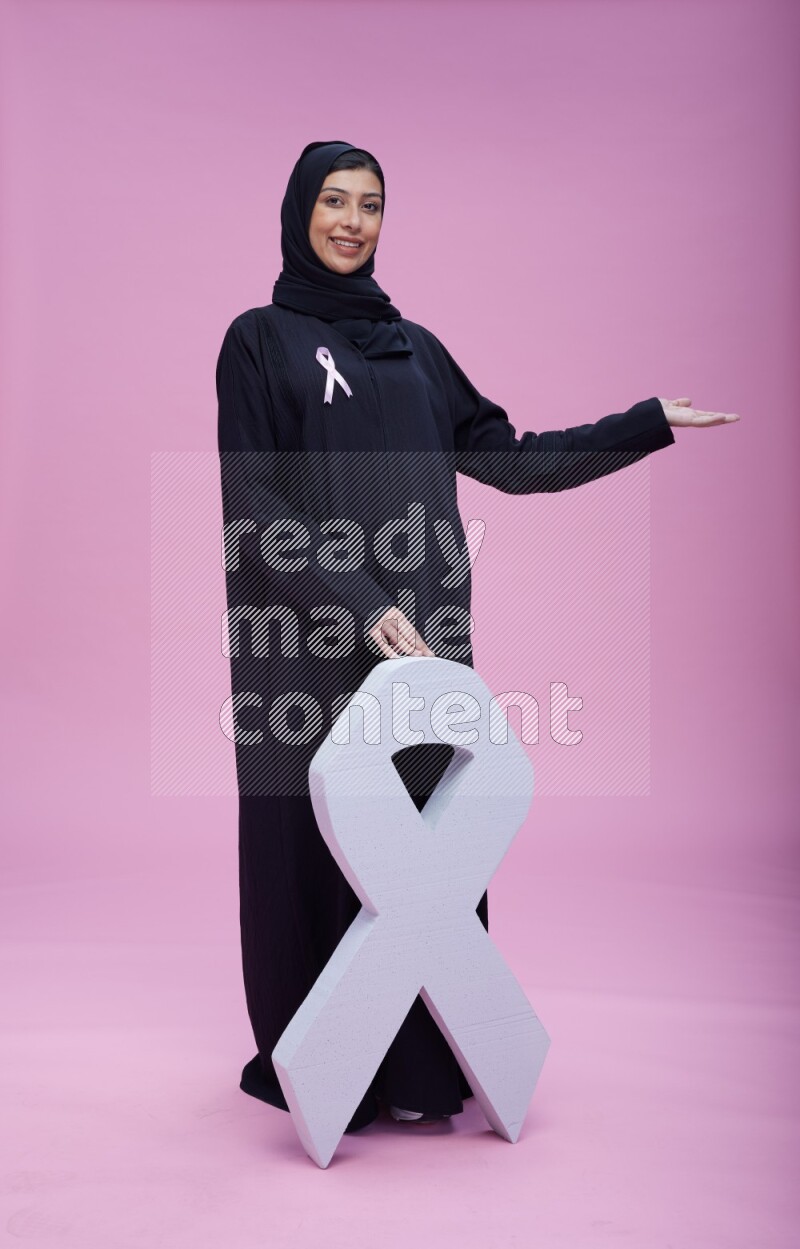Saudi woman wearing pink ribbon on abaya standing holding awareness ribbon on plain pink background