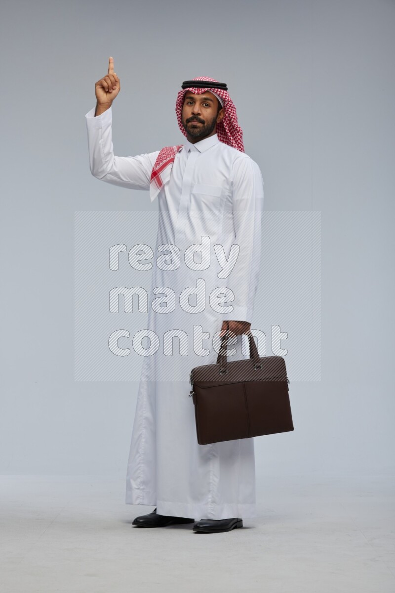 Saudi man Wearing Thob and shomag standing holding bag on Gray background